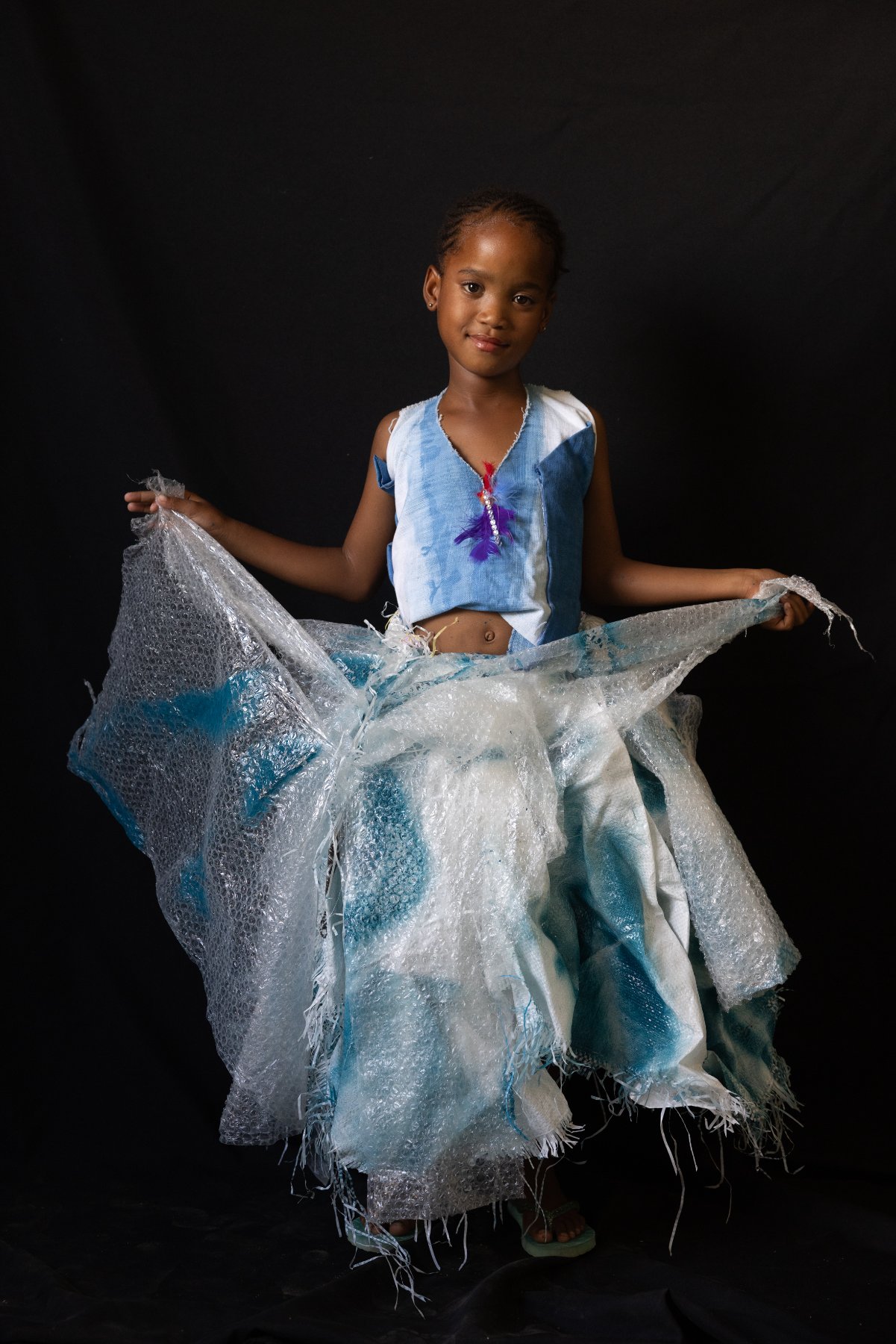 A child in a fringed recycled-material outfit and hat against a black backdrop