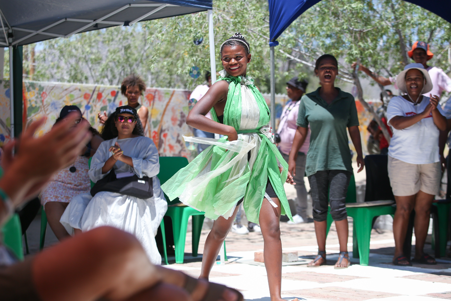 A young performer in a dress made from green and clear plastic bags dances under a gazebo at a community activation, cheered on by the crowd
