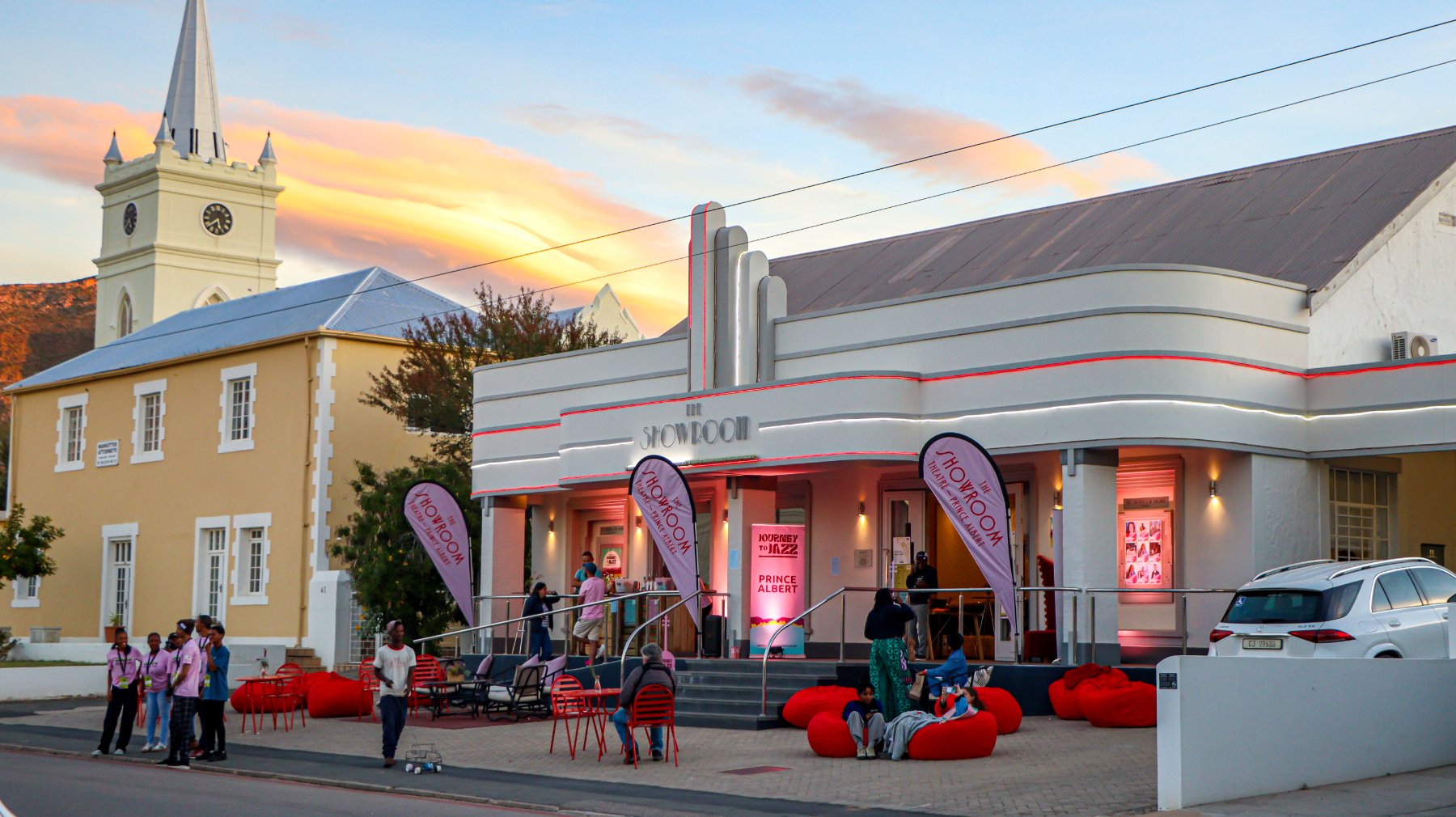 The Showroom theatre at dusk, Art-Deco façade lit in pink and red beside the church clock tower, with people gathered on red bean bags out front